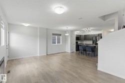 Open concept living area with light wood-style flooring and white walls, featuring a modern kitchen with dark cabinetry, stainless steel appliances, and a breakfast bar - 