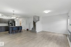 Spacious open-concept living area featuring light-toned flooring, a modern kitchen with dark cabinetry and an island, a staircase with carpeted treads, and a fireplace with a light-colored surround - 