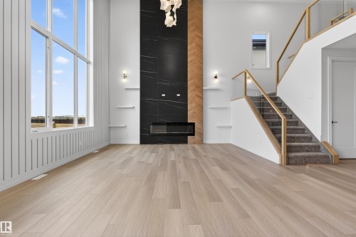 Spacious living area featuring a two-story window array, wood-finish flooring, and a contemporary fireplace with a dark stone-finish surround - 63 Jubilation Drive, St. Albert, AB - Indoor With Fireplace