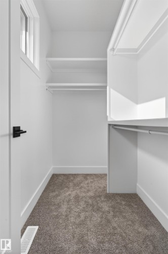 Walk-in closet featuring built-in shelving, hanging rods, an upper transom window, neutral carpeting, and a black lever door handle - 63 Jubilation Drive, St. Albert, AB - Indoor With Storage