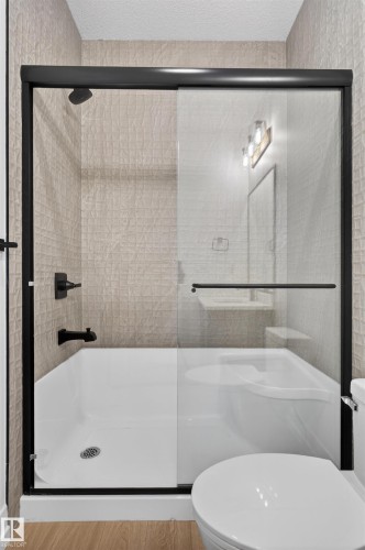 Bathroom featuring a tub/shower combination with black-framed glass enclosure, textured tile surround, matte black fixtures, and wood-finish flooring - 63 Jubilation Drive, St. Albert, AB - Indoor Photo Showing Bathroom