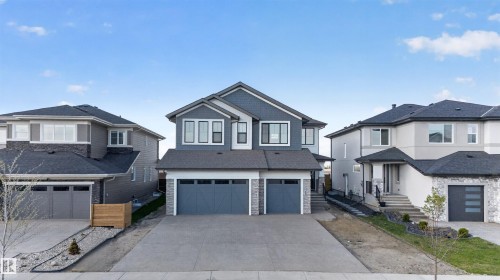 Modern duplex featuring a dark gray facade with white trim, a double attached garage, a spacious concrete driveway, an asphalt shingle roof, and a stone accent foundation - 63 Jubilation Drive, St. Albert, AB - Outdoor With Facade
