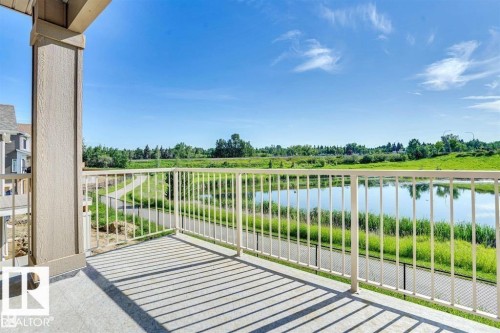 This property features a balcony with a white railing, offering views of a pond, a paved pathway, and lush green landscaping under a clear sky - 47 Rosewood Way, St. Albert, AB - Outdoor With Balcony