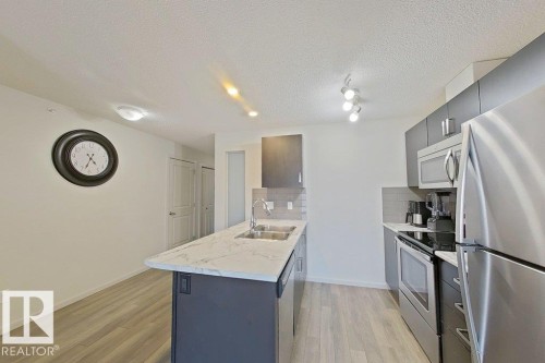 The kitchen features a functional island with a double sink, stainless steel appliances, and sleek grey cabinetry - 429 390 Windermere Rd, Edmonton, AB - Indoor Photo Showing Kitchen With Stainless Steel Kitchen With Double Sink