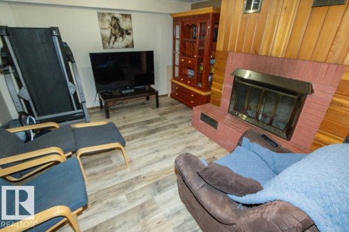 The living area features wood paneled walls, a brick fireplace with a brass firebox, and light-toned flooring - 5016 47 St, Myrnam, AB - Indoor Photo Showing Living Room With Fireplace