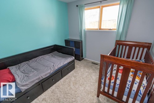 This room features a window with light-colored drapes, gray carpeting, and a wall painted in a vibrant blue hue - 5016 47 St, Myrnam, AB - Indoor Photo Showing Bedroom