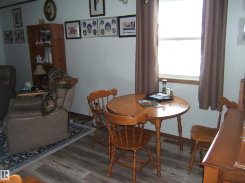 This inviting living area features wood-look flooring, a window with brown curtains, and a wooden dining table with chairs - 4702 52 Avenue, Two Hills, AB - Indoor Photo Showing Dining Room