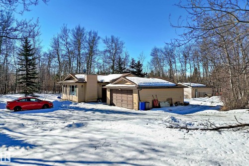 The property features a single-story structure with an attached garage and a detached outbuilding, set amidst a wooded backdrop - 100 23016 Twp Rd 504, Rural Leduc County, AB - Outdoor