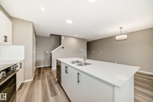 Kitchen featuring a large island with an undermount double sink and stainless steel faucet, white cabinetry with matte black hardware, integrated stainless steel appliances, white subway tile backsplash, and wood-finish flooring - 20415 16 Avenue, Edmonton, AB 