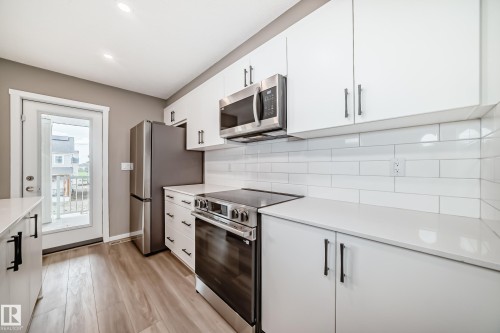 Modern kitchen featuring white cabinetry with black hardware, stainless steel appliances, light countertops, a white subway tile backsplash, and wood-finish flooring - 20415 16 Avenue, Edmonton, AB 