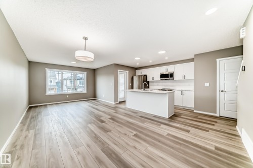 Open-concept living area featuring wood-finish flooring, a white kitchen island, stainless steel appliances, white cabinetry, and recessed lighting - 20415 16 Avenue, Edmonton, AB 