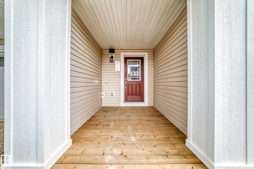 Covered front porch featuring wood-finish flooring, white architectural columns, beige vinyl siding, and a red entry door with decorative glass - 20415 16 Avenue, Edmonton, AB 