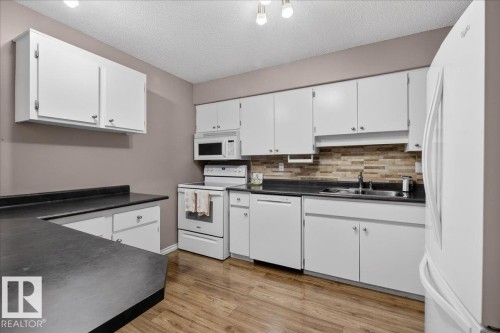 The kitchen features white cabinetry, dark countertops, a tiled backsplash, and wood-style flooring - 3240 130A Avenue, Edmonton, AB - Indoor Photo Showing Kitchen With Double Sink