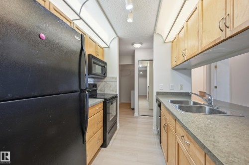 The kitchen features light-toned wooden cabinetry, a double basin stainless steel sink, and a black refrigerator, oven, and microwave - 314 2430 Guardian Road, Edmonton, AB - Indoor Photo Showing Kitchen With Double Sink