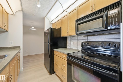 The kitchen features light wood cabinetry, stone countertops, and black appliances including a refrigerator, oven, and microwave - 314 2430 Guardian Road, Edmonton, AB - Indoor Photo Showing Kitchen