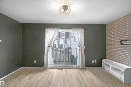 Living area featuring light wood flooring, a decorative ceiling light fixture, a sliding glass door with a balcony, and an accent wall with a brick pattern - 13 3751 12 Street, Edmonton, AB - Indoor Photo Showing Other Room