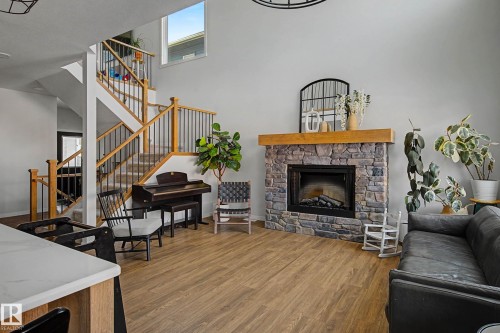 This living space features a stone fireplace with a wooden mantel, a staircase with wooden handrails and black balusters, and light wood-look flooring - 34 Riverhill Crescent, St. Albert, AB - Indoor Photo Showing Living Room With Fireplace