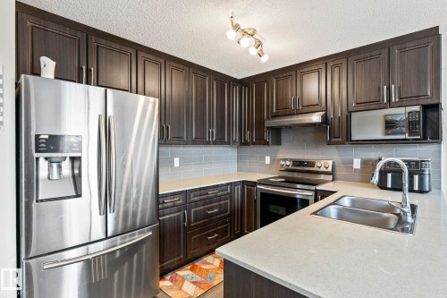 The kitchen features dark wood cabinetry, stainless steel appliances, light-colored countertops, and a subway tile backsplash - 483 Ebbers Way Nw, Edmonton, AB - Indoor Photo Showing Kitchen With Double Sink With Upgraded Kitchen
