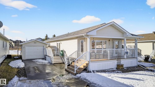 The property features a single-story residence with light-colored siding, a covered porch with white railings, and an attached single-car garage with a driveway - 1727 Jubilee Loop, Sherwood Park, AB - Outdoor With Deck Patio Veranda