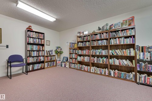 This library area features extensive shelving filled with books, a purple chair, and carpeted flooring - 304 17151 94A Avenue, Edmonton, AB - Indoor Photo Showing Other Room