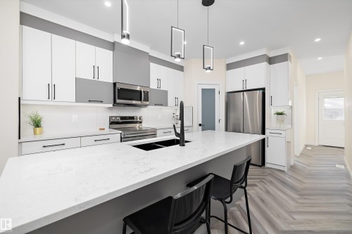 The kitchen features an island with a light-colored countertop and a dark sink, modern pendant lighting, white and gray cabinetry, and stainless steel appliances including a microwave, oven, and refrigerator - 8321 81Ave, Edmonton, AB - Indoor Photo Showing Kitchen With Double Sink With Upgraded Kitchen