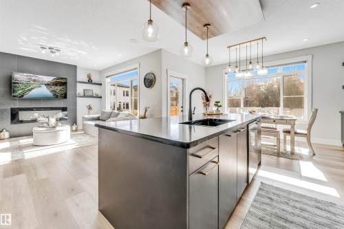 Open concept living area featuring light-colored hardwood floors, a kitchen island with a dark countertop and black sink, and a modern fireplace with a dark stone surround - 1531 151 Avenue, Edmonton, AB - Indoor Photo Showing Kitchen With Upgraded Kitchen