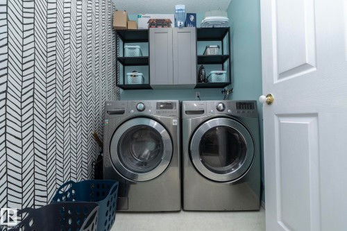 Dedicated laundry area featuring modern front-loading washer and dryer units, a wall-mounted cabinet with open shelving, and a patterned accent wall - 6 Glaewyn Estates, St. Albert, AB - Indoor Photo Showing Laundry Room
