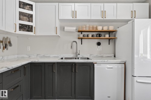 The kitchen features white upper cabinetry with gold hardware, dark lower cabinetry with dark hardware, a stainless steel sink with a gooseneck faucet, and white countertops with grey veining - 1873 151 Avenue, Edmonton, AB - Indoor Photo Showing Kitchen With Double Sink