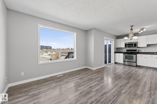 The property features an open-concept living area with wood-look flooring, a large window providing views of the surrounding neighborhood, and a kitchen equipped with white cabinetry, dark countertops, and stainless steel appliances - 30 450 Hyndman Crescent, Edmonton, AB - Indoor Photo Showing Kitchen