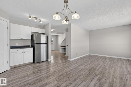 The open concept living area features light-colored walls and wood-look flooring, with a kitchen area offering white cabinetry, dark countertops, and a stainless steel refrigerator - 30 450 Hyndman Crescent, Edmonton, AB - Indoor Photo Showing Kitchen