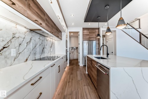 Modern kitchen featuring white cabinetry, wood-grain upper cabinets, a stone backsplash, and an island with an integrated sink and pendant lighting - 7665 Kimiwan Crescent, Edmonton, AB - Indoor Photo Showing Kitchen With Upgraded Kitchen