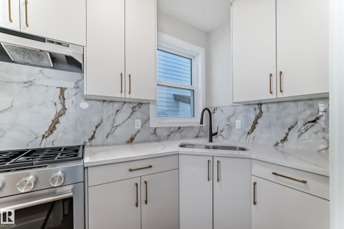 The kitchen features white cabinetry with gold hardware, a white and grey marble-patterned backsplash, a stainless steel range with a matching range hood, and a corner sink with a black faucet - 7665 Kimiwan Crescent, Edmonton, AB - Indoor Photo Showing Kitchen With Double Sink With Upgraded Kitchen