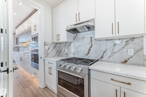 The kitchen features white cabinetry with gold-toned hardware, a stainless steel range with a ventilation hood, and marble-patterned backsplash and countertops - 7665 Kimiwan Crescent, Edmonton, AB - Indoor Photo Showing Kitchen With Upgraded Kitchen