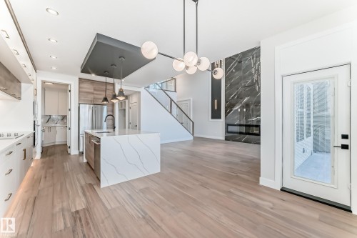 This open-concept property features hardwood flooring, a modern kitchen island with an integrated sink and pendant lighting, and a striking black marble fireplace - 7665 Kimiwan Crescent, Edmonton, AB - Indoor Photo Showing Kitchen With Upgraded Kitchen
