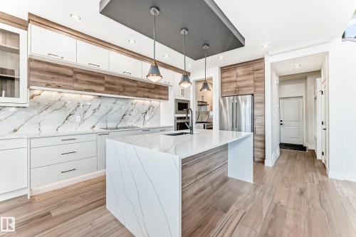 The kitchen features a large island with a waterfall countertop, white cabinetry with wood accents, and a full-height marble-style backsplash - 7665 Kimiwan Crescent, Edmonton, AB - Indoor Photo Showing Kitchen With Upgraded Kitchen