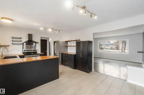 The kitchen features a large island with a wooden countertop and a sink with a gold-toned faucet, stainless steel appliances including a range and refrigerator, black cabinetry, and light-toned floor tiles - 12121 38 Street, Edmonton, AB - Indoor Photo Showing Kitchen With Double Sink