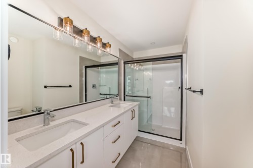 This bathroom features a double vanity with an expansive mirror, a contemporary light fixture, and a glass-enclosed shower with a black frame - 10718 149 Street Nw, Edmonton, AB - Indoor Photo Showing Bathroom