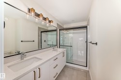 This bathroom features a double vanity with an expansive mirror, a contemporary light fixture, and a glass-enclosed shower with a black frame - 