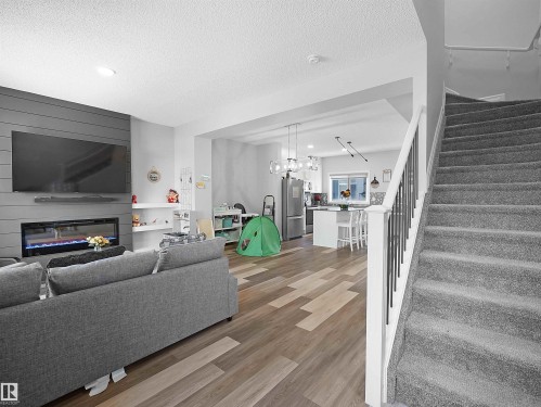 Open concept living space featuring a modern fireplace with a horizontally paneled accent wall, wood plank style flooring, and a carpeted staircase with a white and black railing - 138 804 Welsh Drive, Edmonton, AB - Indoor Photo Showing Living Room With Fireplace