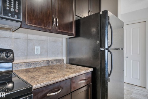 The kitchen features dark wood cabinetry, a tiled backsplash, and a black refrigerator - 108 1204 156 Street, Edmonton, AB - Indoor Photo Showing Kitchen