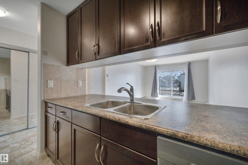 The kitchen features dark wood cabinetry, a double basin stainless steel sink with a chrome faucet, and a stone-patterned countertop - 108 1204 156 Street, Edmonton, AB - Indoor Photo Showing Kitchen With Double Sink