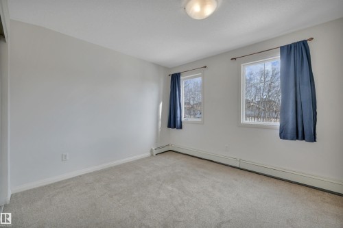 The room features light gray carpeting, neutral white walls, and two windows with blue curtains - 108 1204 156 Street, Edmonton, AB - Indoor Photo Showing Other Room