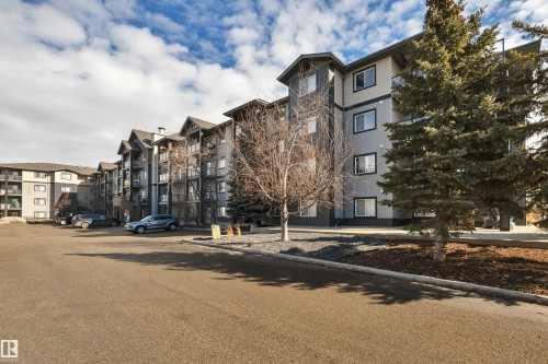 Exterior view of the property, featuring a multi-story building with balconies, a paved parking area, and landscaping with trees and wood chips - 108 1204 156 Street, Edmonton, AB - Outdoor With Facade