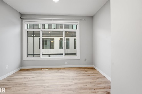 This bright room features light wood-style flooring, white baseboards, and a large window providing views of nearby modern architecture - 40 1729 Keene Crescent, Edmonton, AB - Indoor Photo Showing Other Room