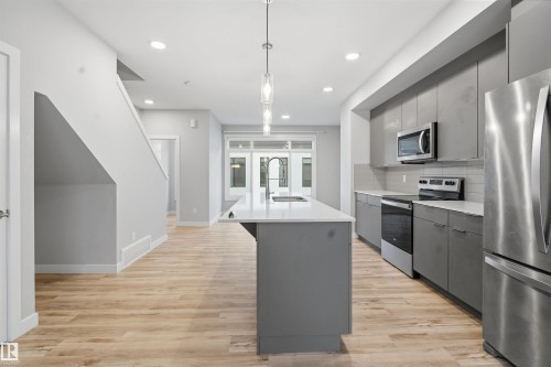 The kitchen features an island with a sink and white countertop, grey cabinetry, and stainless steel appliances including a refrigerator, microwave, and oven - 40 1729 Keene Crescent, Edmonton, AB - Indoor Photo Showing Kitchen With Upgraded Kitchen