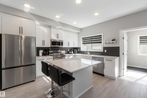 The kitchen features a large island with a countertop, stainless steel appliances, white cabinetry with dark hardware, a dark tiled backsplash, and light-colored flooring - 29 Gambel Loop, Spruce Grove, AB - Indoor Photo Showing Kitchen With Upgraded Kitchen