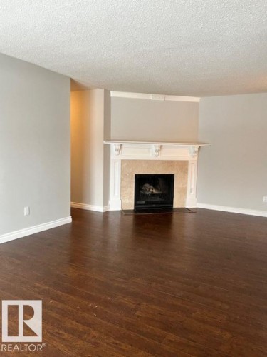 This room features dark wood flooring, light-colored walls, and a fireplace with a decorative mantel and stone surround - 4 10836 116 Street, Edmonton, AB - Indoor Photo Showing Living Room With Fireplace