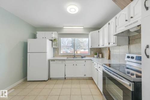 The kitchen features white cabinetry with dark hardware, tile flooring, a window above the sink, and white appliances including a refrigerator, dishwasher, and stove - 15704 91 Avenue, Edmonton, AB - Indoor Photo Showing Kitchen With Double Sink