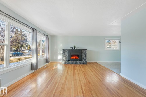 This inviting living space features light wood flooring, large windows providing natural light, and a fireplace with a stone surround - 15704 91 Avenue, Edmonton, AB - Indoor Photo Showing Living Room With Fireplace