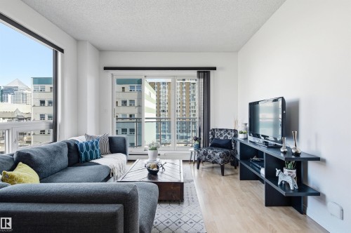 The living area features light-colored flooring, a large window providing views of surrounding buildings, and a glass sliding door opening to an outdoor balcony - 601 10504 99 Avenue, Edmonton, AB - Indoor Photo Showing Living Room